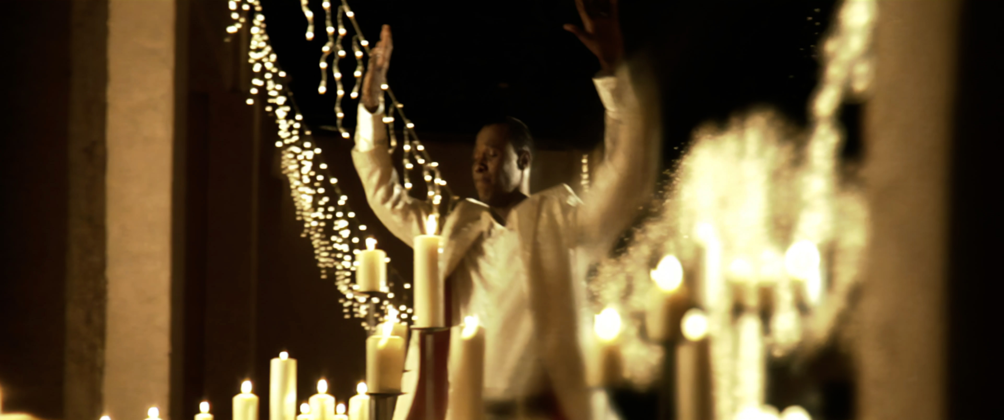 Micah Stampley with candles and string lights, arms raised. I Believe